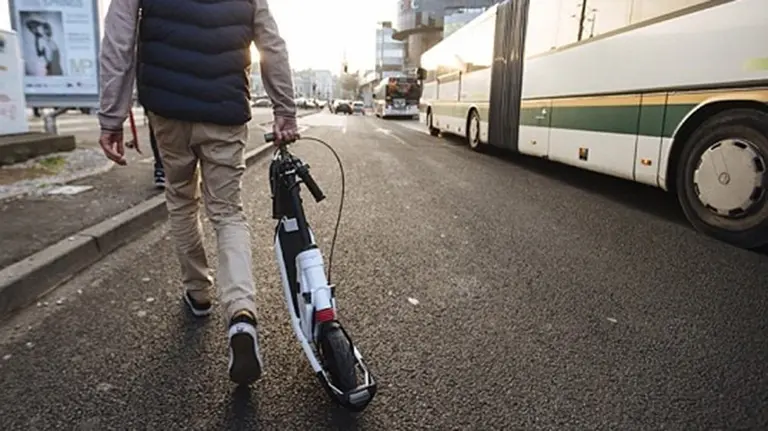 Imagen de un conductor de un patinete el&eacute;ctrico caminando por la calle junto a un autob&uacute;s urbano. ARCHIVO
