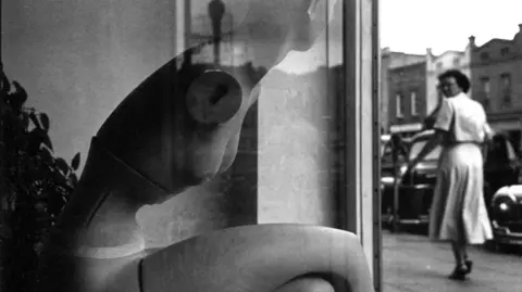 Una mujer camina por la calle frente a un escaparate con un maniquí sin brazos. ELLIOT ERWITT
