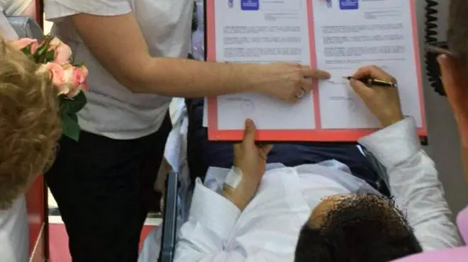 El novio dentro de la ambulancia firmando el acta matrimonial a la concejala del distrito. A la izquierda, se ve el ramo de la novia. Foto AYUNTAMIENTO DE MADRID.