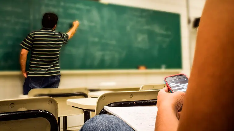 Un profesor da clases mientras un alumno mira el móvil