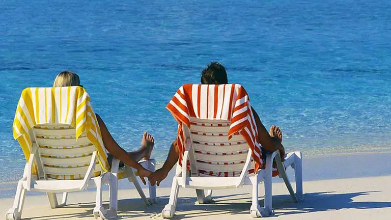 Imagen de una pareja de turistas agarrándose de la mano mientras toman el sol en unas tumbonas en la playa. ARCHIVO