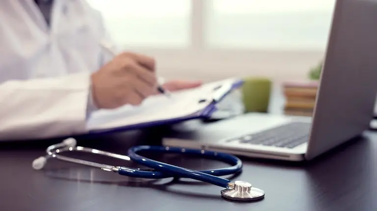 Un médico trabaja con un estetoscopio en la consulta de un centro de salud mientras los pacientes esperan en la sala del ambulatorio ARCHIVO