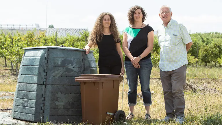 Raquel Igoa Linares (Cafetería-Comedor de El Sario) y los investigadores de la UPNA Amaia Oreja Ostiz y Julio Muro Erreguerena posan en la Finca de Prácticas e Investigación Agrícola, con la compostadora. CEDIDA