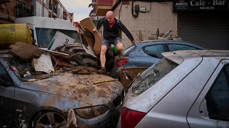 Un hombre pasa por encima de varios coches apilados para cruzar una calle de Alfafar. PABLO LASAOSA