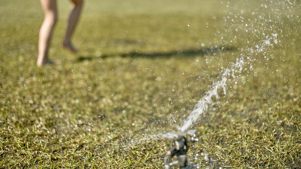 Imagen de un aspersor de un jardín regando el césped mientras una niña se remoja jugando con el agua ARCHIVO