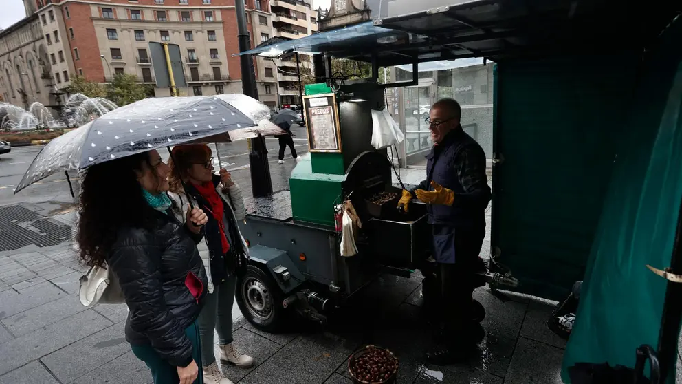 Dos mujeres se resguardan con sus paraguas de la lluvia mientras disfrutan de la tradición otoñal de comprar castañas en un puesto situado en la Avenida Carlos III.  La Agencia Estatal de Meteorología anuncia lluvias, más persistentes en el centro y sur de la Comunidad Foral, que podrían arrastrar algo de barro, con alguna tormenta ocasional. Predominarán los cielos nubosos o cubiertos en una jornada en la que el viento soplará flojo variable y las temperaturas mínimas experimentarán un ligero ascenso en la Ribera y pocos cambios en el resto y las máximas apenas sufrirán variaciones. EFE/ Jesus Diges